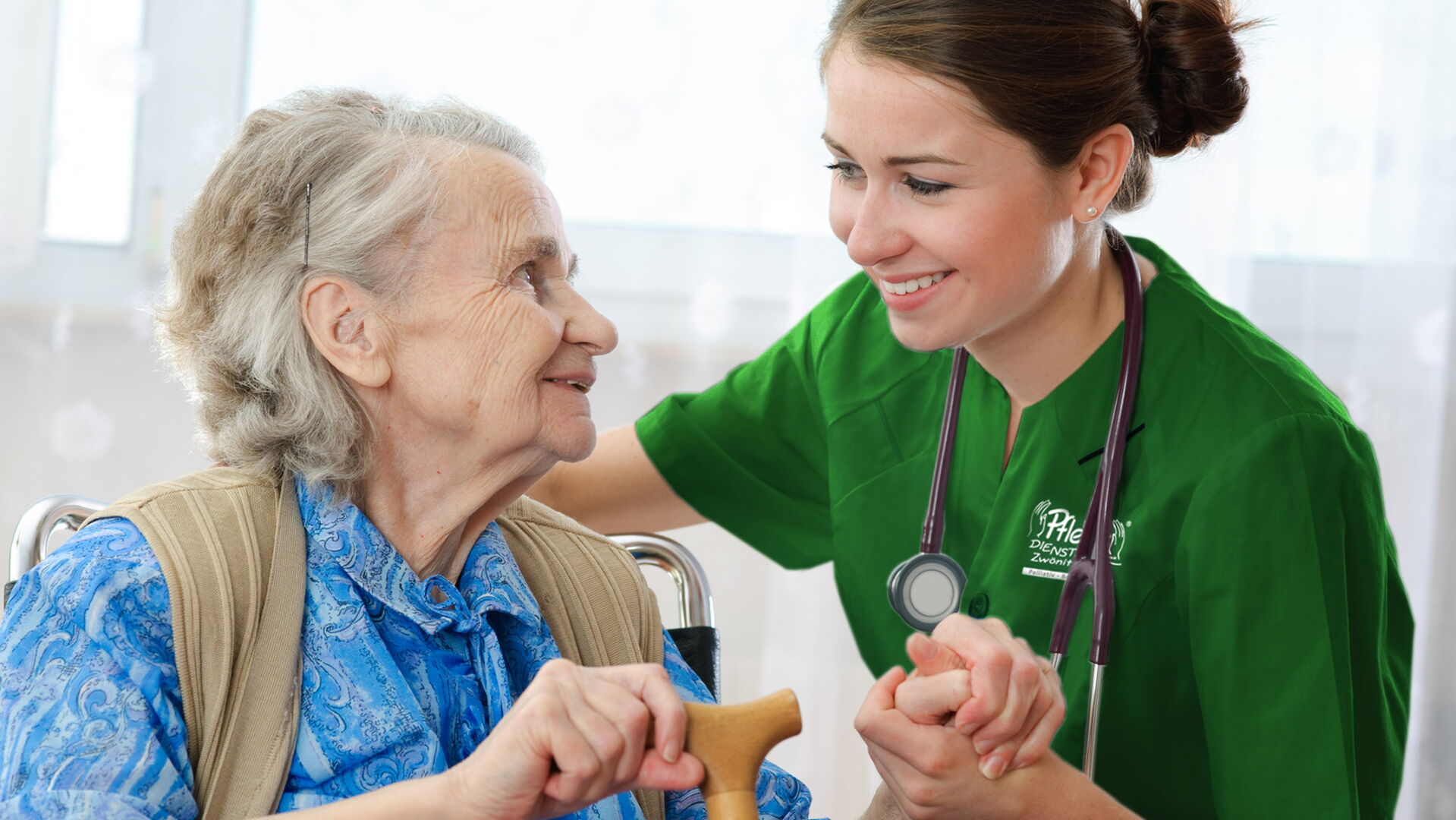 Pflegekraft in grüner Dienstkleidung hält lächelnd die Hand einer Seniorin im Rollstuhl