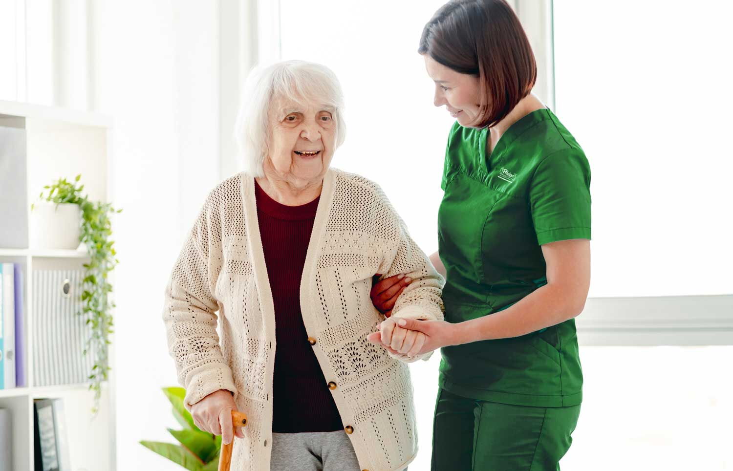 Pflegekraft in grüner Dienstkleidung stützt eine Seniorin beim Gehen in ihrer Wohnung
