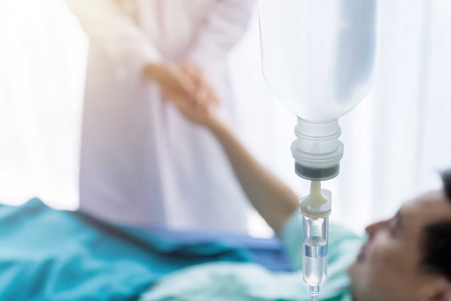 Infusionsflasche am Krankenbett, im Hintergrund eine Pflegekraft bei einem liegenden Patienten